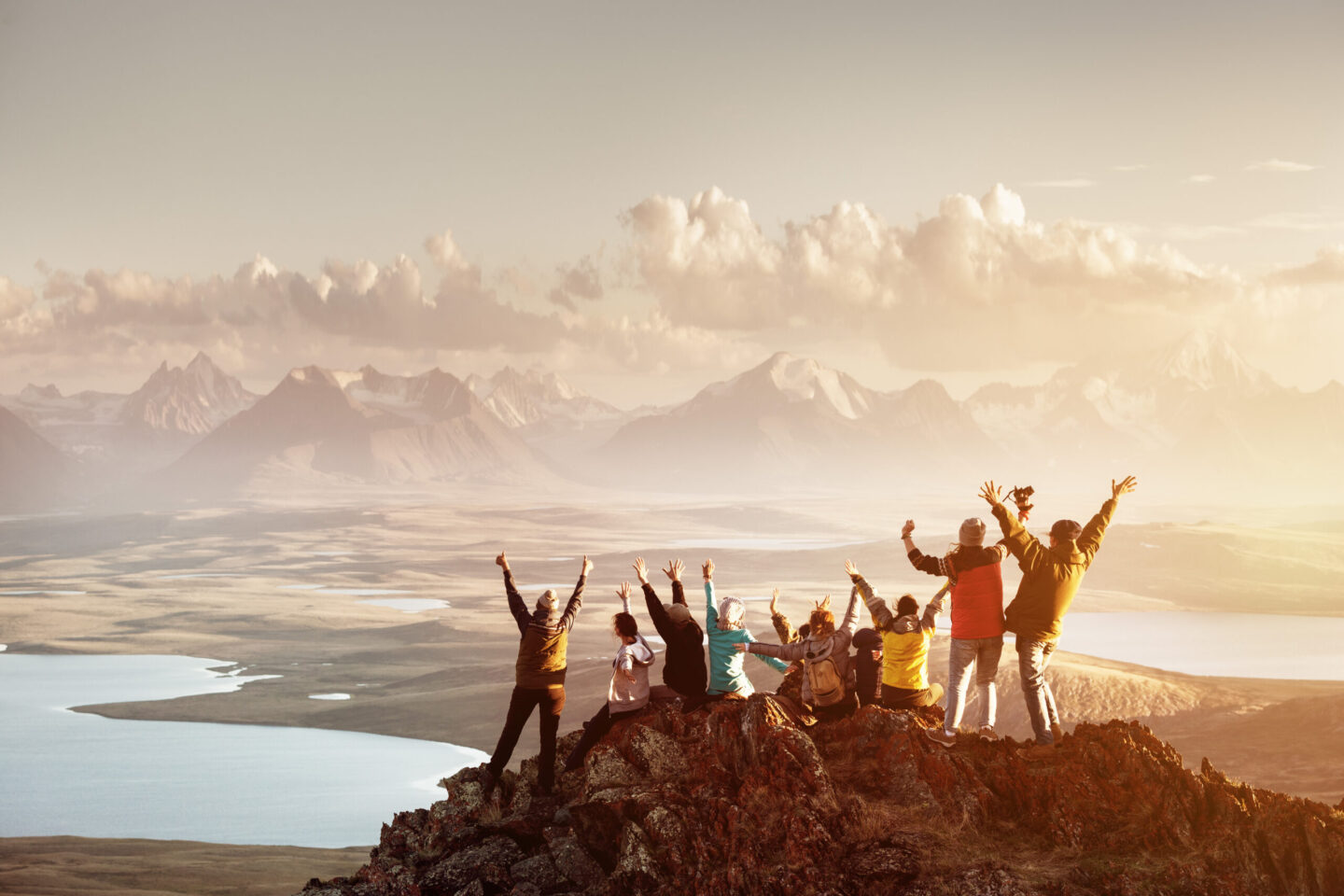 A group of people sitting on a mountain peak, raising their arms in celebration, with a vast landscape and mountains in the distance under a cloudy sky.
