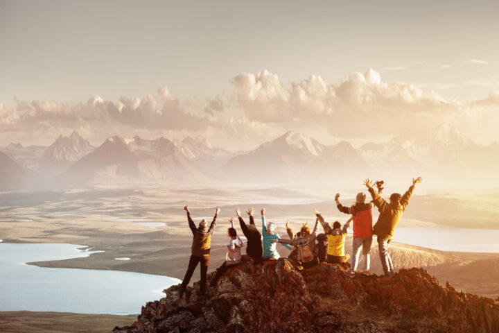 A group of people sitting on a mountain peak, raising their arms in celebration, with a vast landscape and mountains in the distance under a cloudy sky.