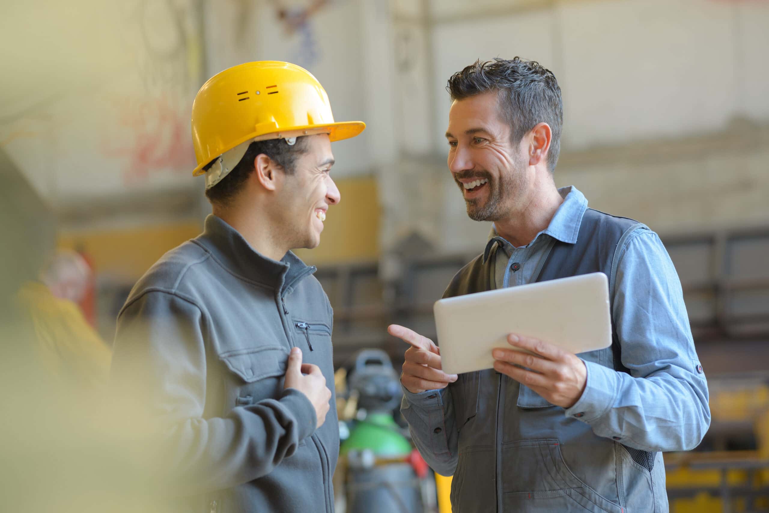 Two men in work attire smiling, one wearing a yellow hard hat, the other holding a tablet in a construction setting.