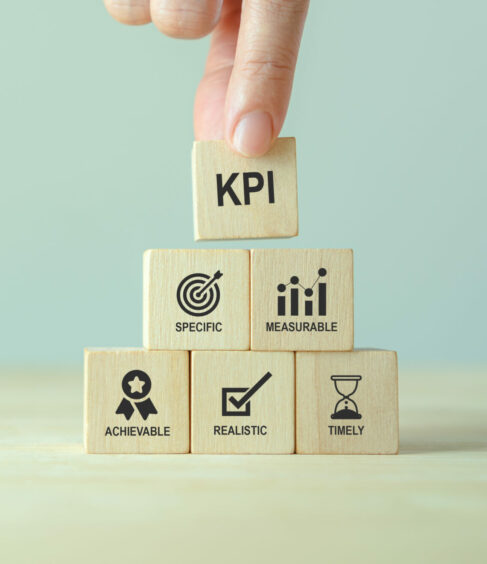 A hand places a wooden block labelled "KPI" on top of a pyramid of blocks. The lower blocks show icons and words: "Specific," "Measurable," "Achievable," "Realistic," and "Timely."