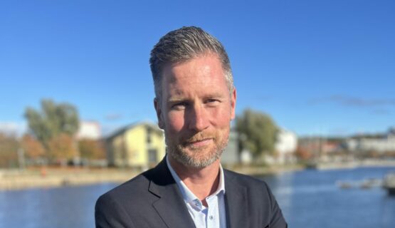 A person in a suit stands by a waterfront with buildings and trees in the background under a clear blue sky.