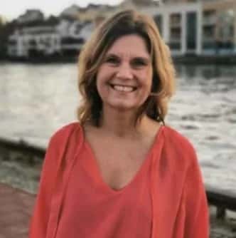 A woman smiling, wearing a coral top, standing in front of a body of water with buildings in the background.