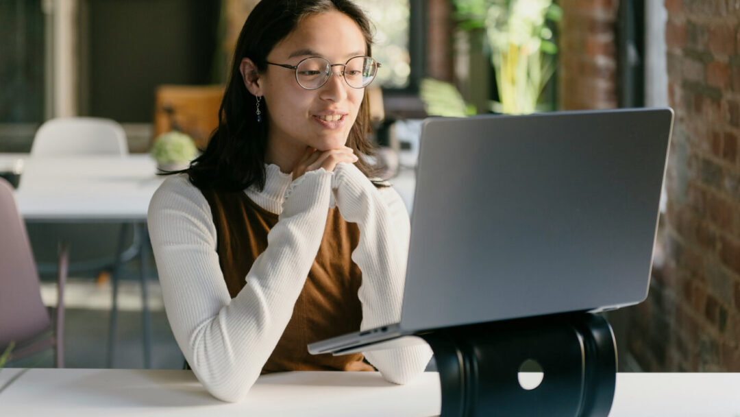 A person with glasses, wearing a white sweater and brown top, is sitting at a desk and looking at a laptop. The background shows a modern, well-lit room with brick walls and plants.