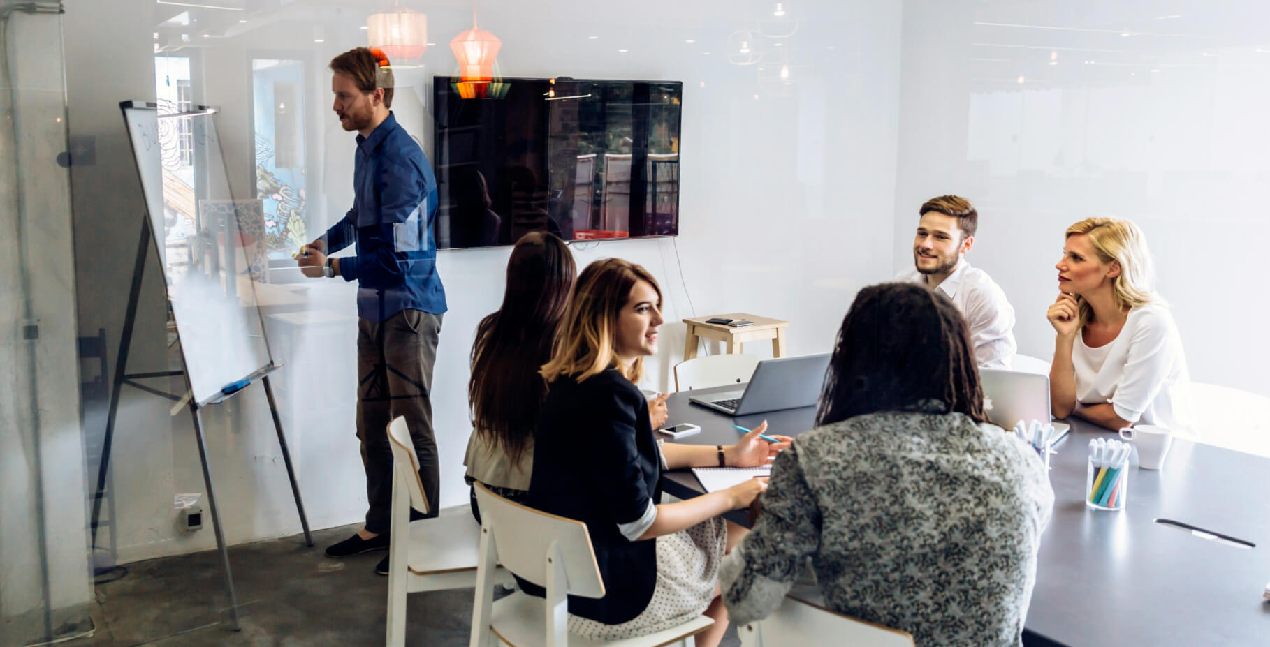 A group of people in a modern office setting, sitting around a table with a laptop. One person stands at a flipchart, presenting ideas.