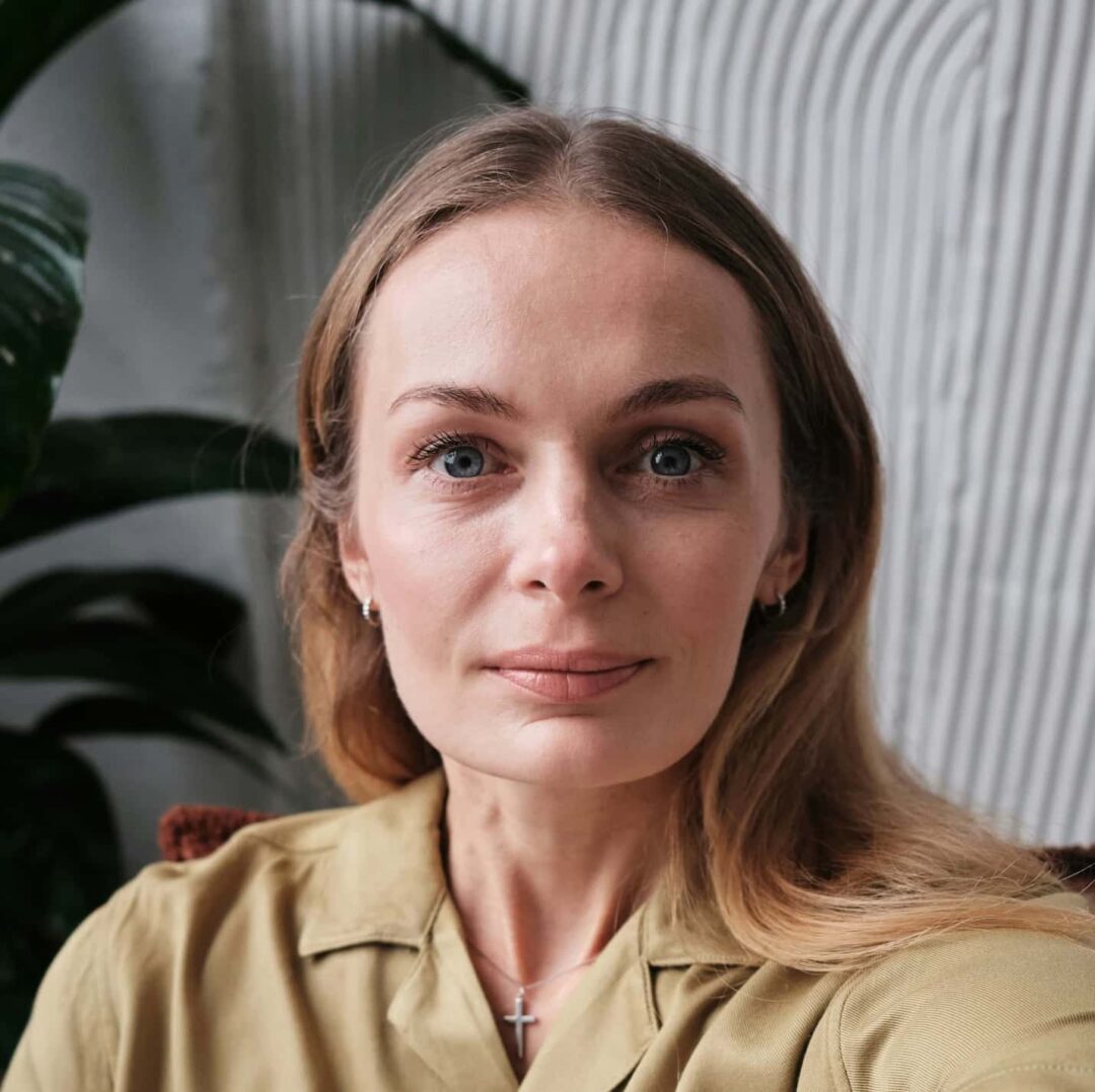 A woman with long blonde hair and blue eyes wearing a light brown shirt and a cross necklace, sitting indoors with a plant in the background.