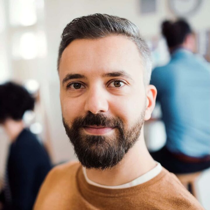 A man with a beard and short hair smiling, wearing a brown jumper, in an indoor setting with blurred people in the background.
