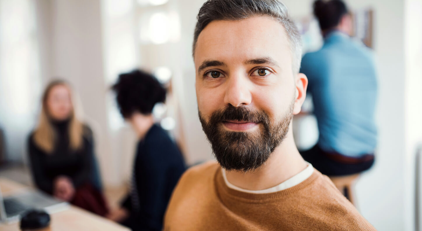 A man with a beard smiles at the camera in an office environment. In the background, three people are engaged in conversation, seated around a table with a laptop. The image is softly focused on the man.