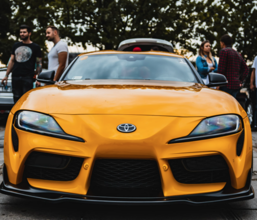 Yellow Toyota sports car parked in front, with a group of people standing nearby. Trees are visible in the background.