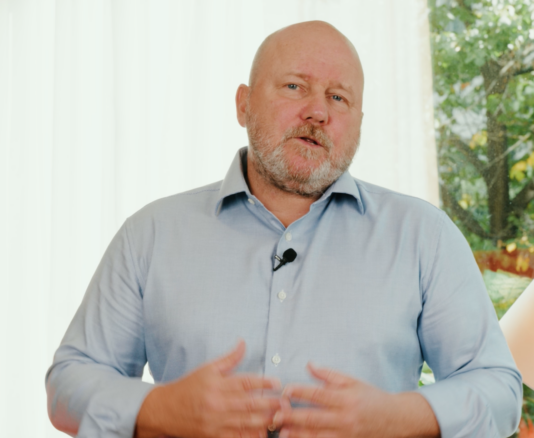 A man with a beard wearing a light blue shirt stands indoors, speaking, with a microphone clipped to his shirt. A white curtain and a view of greenery are in the background.